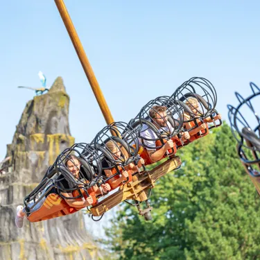 Besucher schweben mit der Attraktion "Hicks Himmelstürmer" im Heide Park Resort liegend durch die Lüfte. Im Hintergrund sind Bäume und ein Fels zu sehen.