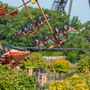 Besucher schweben mit der Attraktion "Hicks Himmelstürmer" im Heide Park Resort liegend durch die Lüfte. Im Hintergrund sind Bäume zu sehen und die Attraktion KRAKE