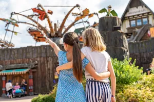 Zwei Mädchen haben die Arme umeinander gelegt. Sie zeigen und schauen in die Ferne. Dort ist "Hicks Himmelsstürmer" zu sehen. Eine Attraktion im Heide Park Resort. Besucher schweben liegend durch die Luft
