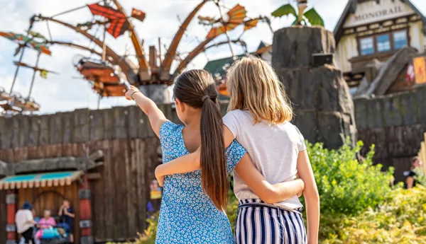 Zwei Mädchen haben die Arme umeinander gelegt. Sie zeigen und schauen in die Ferne. Dort ist "Hicks Himmelsstürmer" zu sehen. Eine Attraktion im Heide Park Resort. Besucher schweben liegend durch die Luft